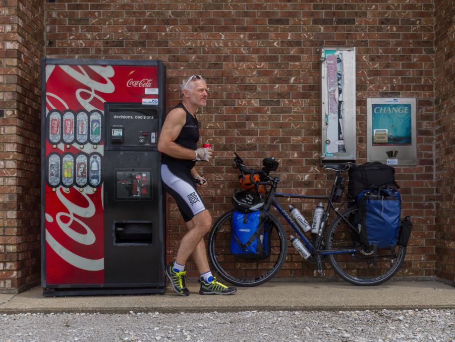Ein Mann in Sportkleidung steht lässig neben einem Fahrrad, das an eine rote Coca-Cola Automatenwand lehnt.