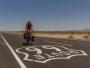 Ein Radfahrer fährt auf einer weiten Straße, die das Logo von Route 66 trägt, unter einem klaren blauen Himmel.